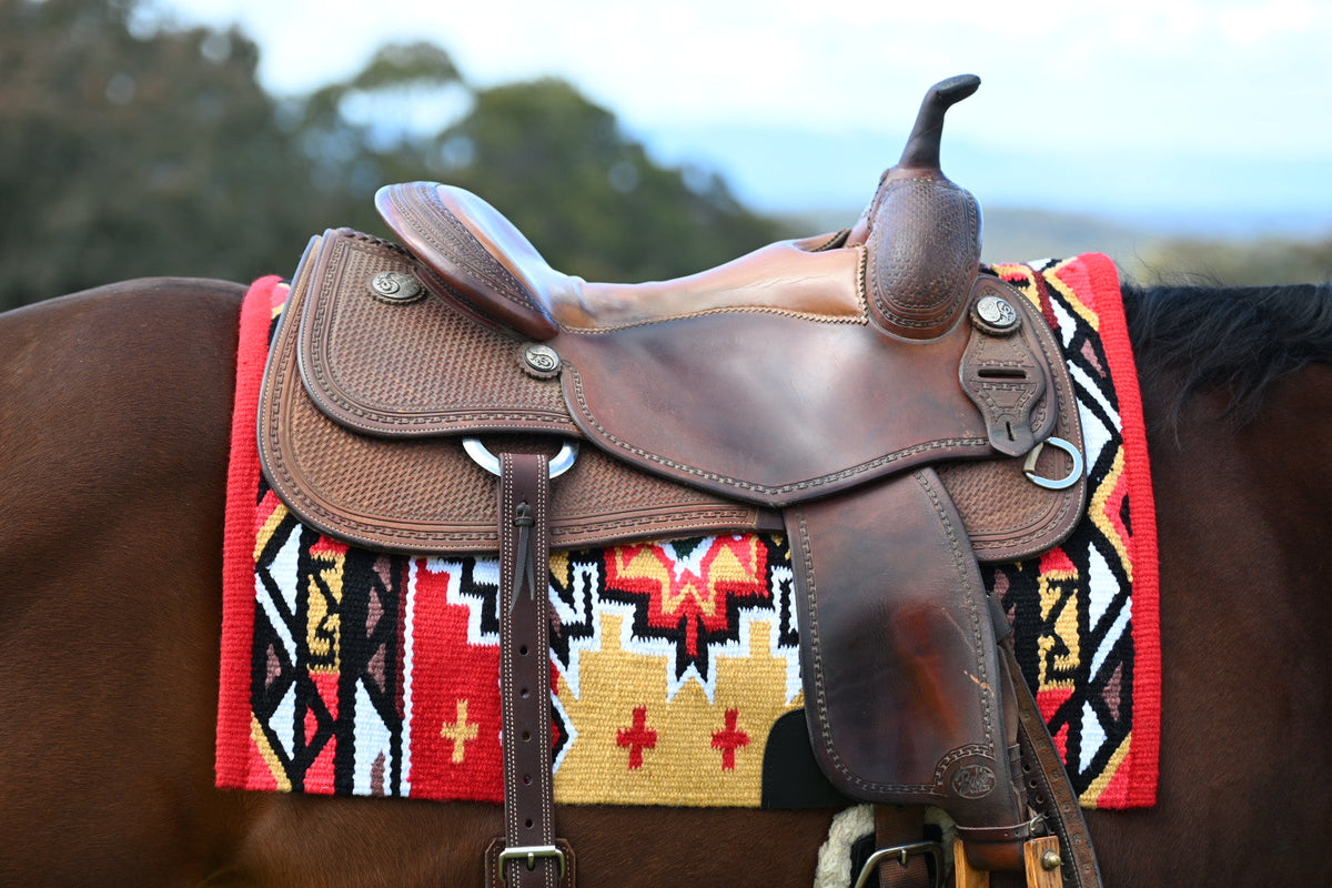 A brown leather western saddle resting on a horse, secured with a red, black, and yellow patterned blanket.