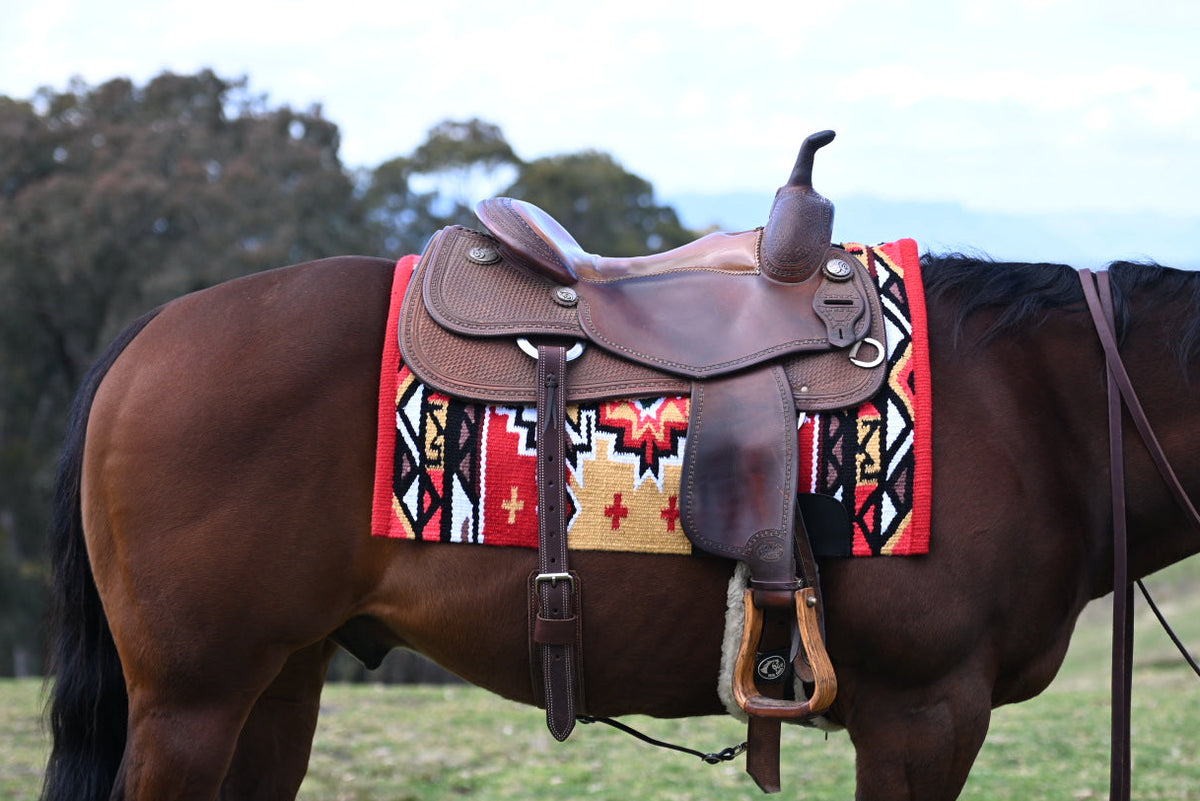 A brown horse standing in a grassy field outdoors, shown in profile with a brown leather western saddle and a colorful red, black, and beige patterned RANCH saddle blanket.